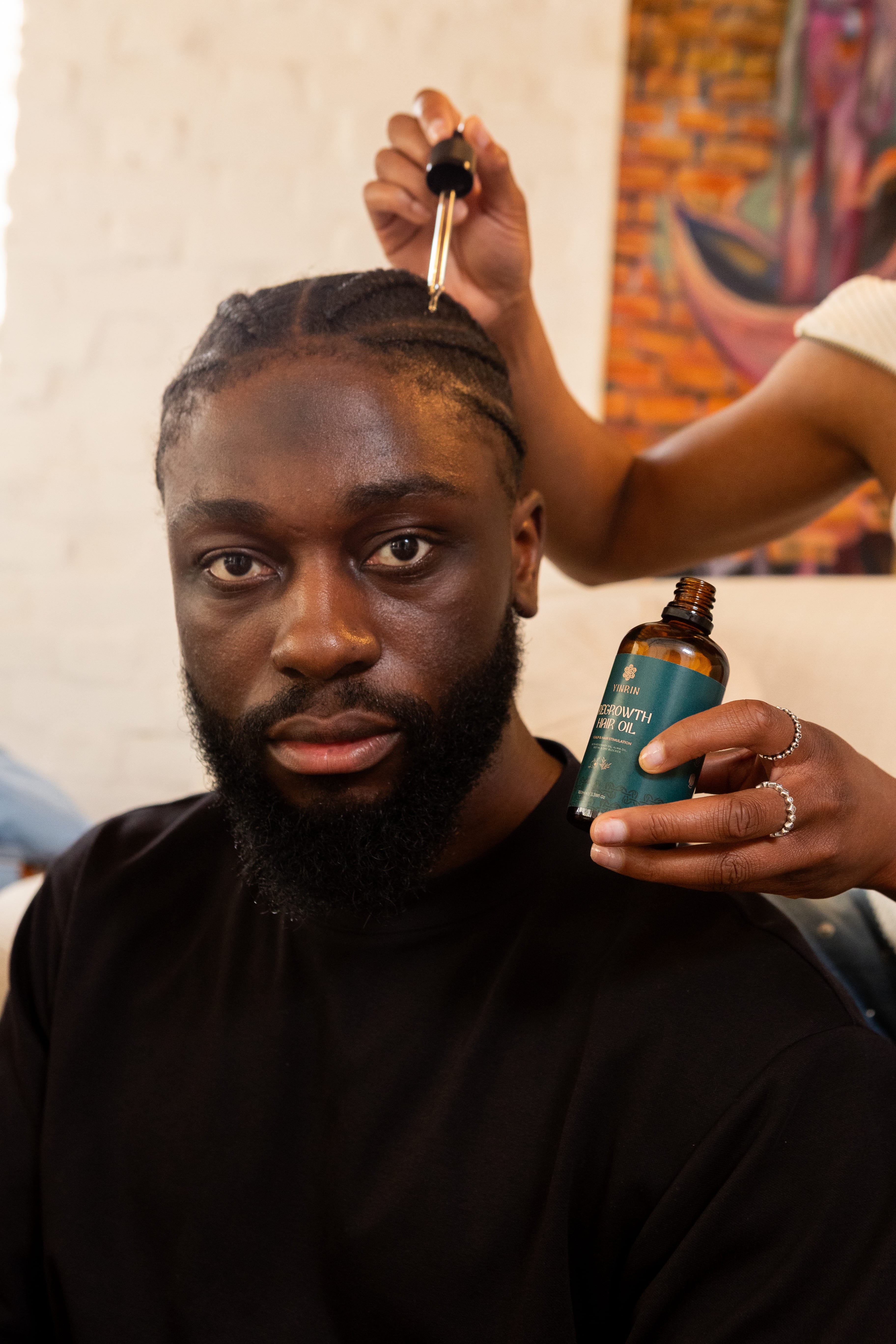 Man with a beard holding a bottle of hair care product, with another person applying it to his hair.